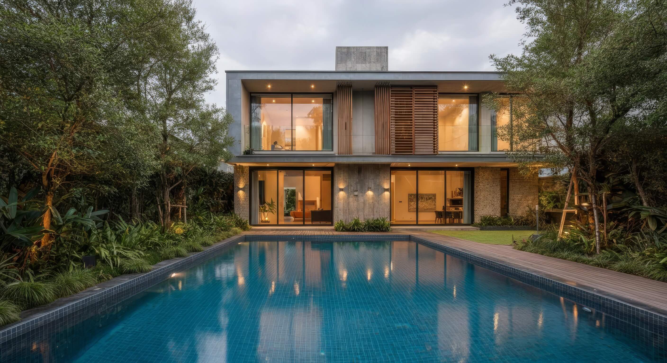 Modern two-story house with large windows and warm interior lighting, reflected in a rectangular swimming pool surrounded by trees and greenery.